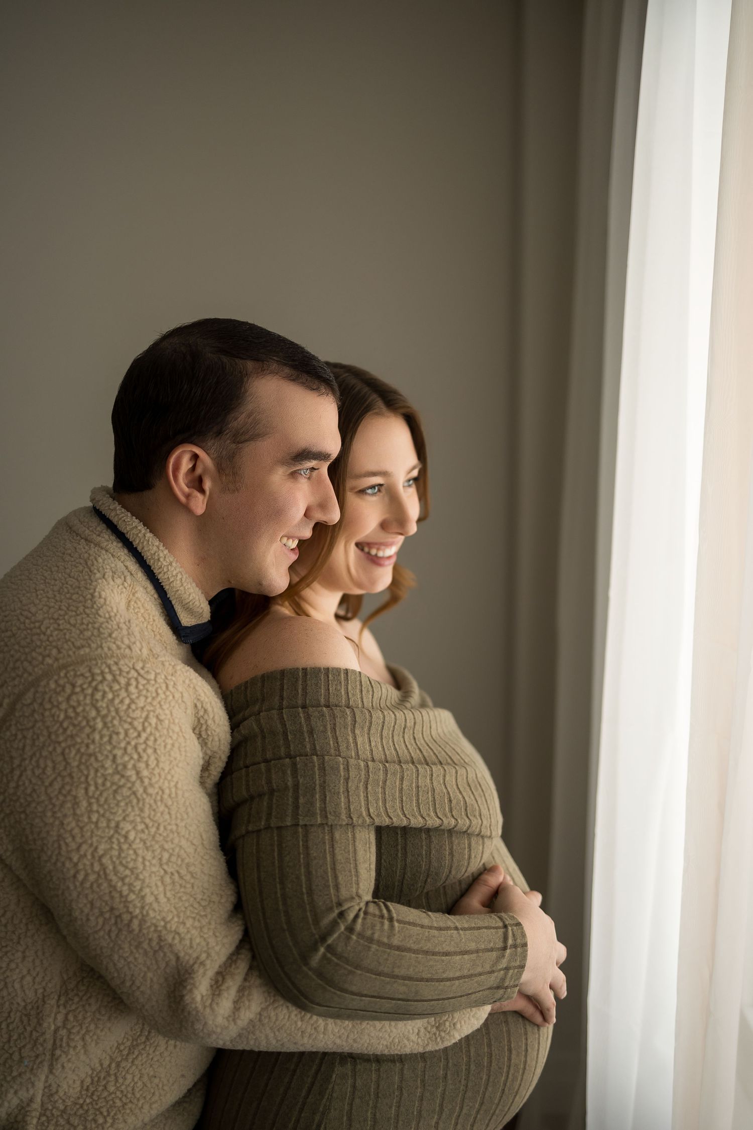 An intimate maternity portrait of a couple embracing by a window in soft natural lighting.