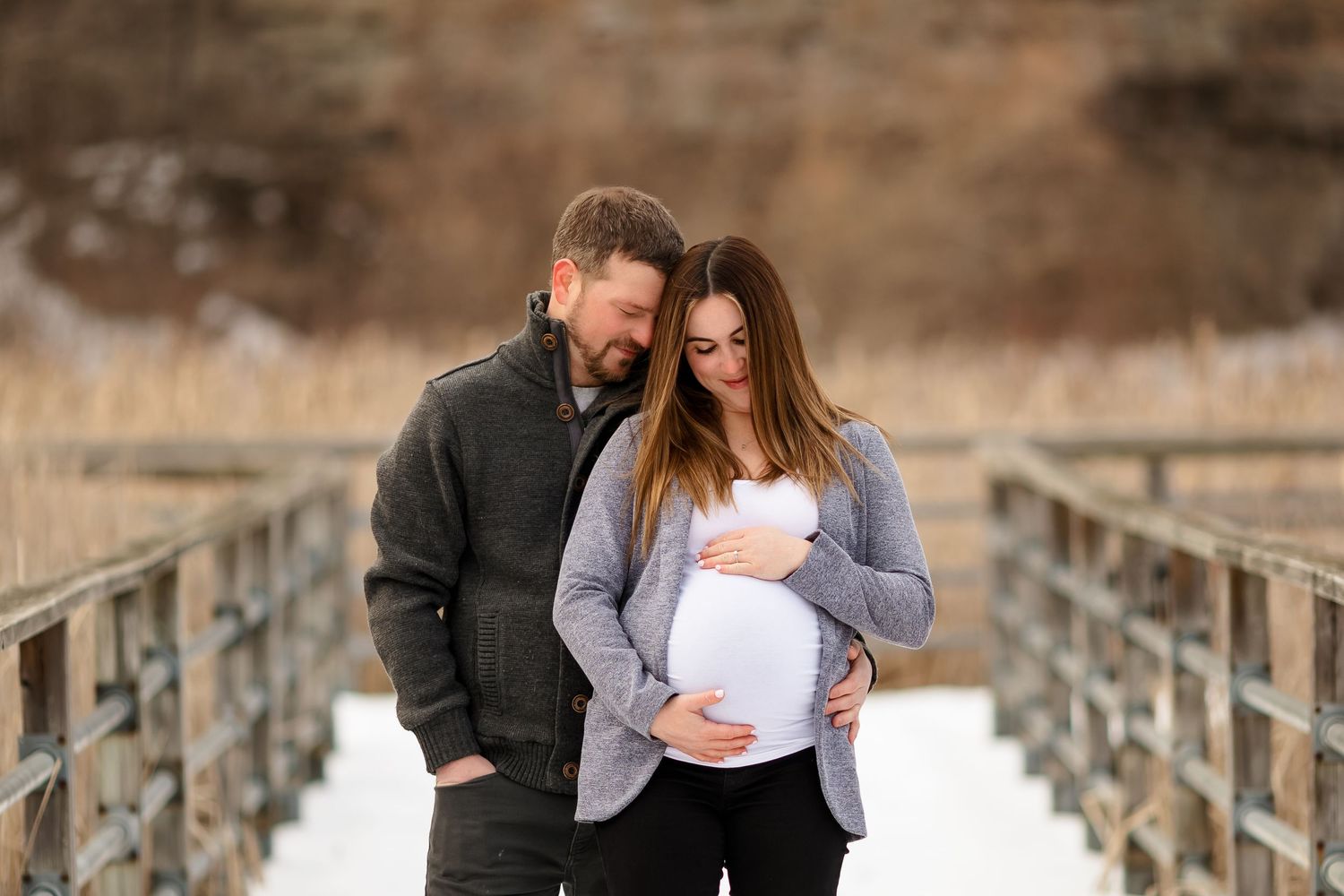 Couple shares intimate moment on snowy bridge during outdoor winter maternity shoot.