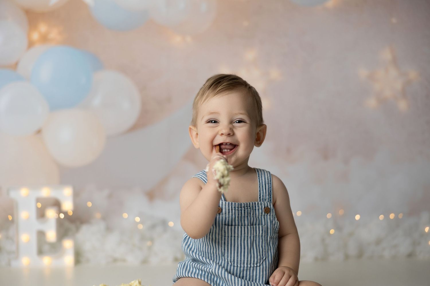 Baby in blue outfit laughing while sitting on white bench surrounded by blue and white balloons.