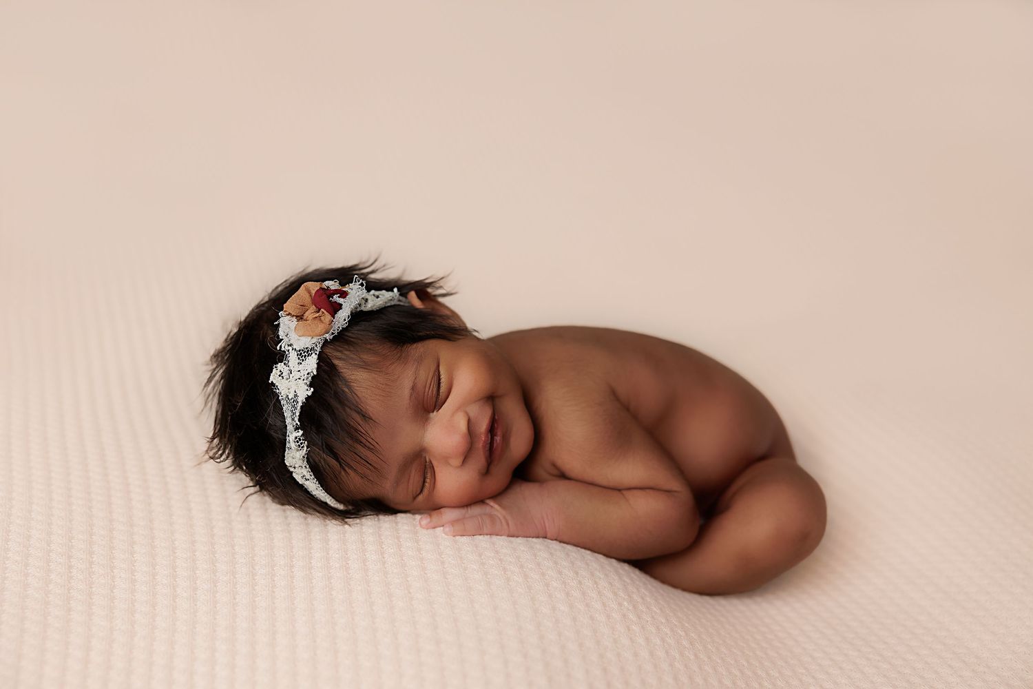 Sleeping newborn wearing delicate silver headband curled up on soft beige backdrop during professional photo session.