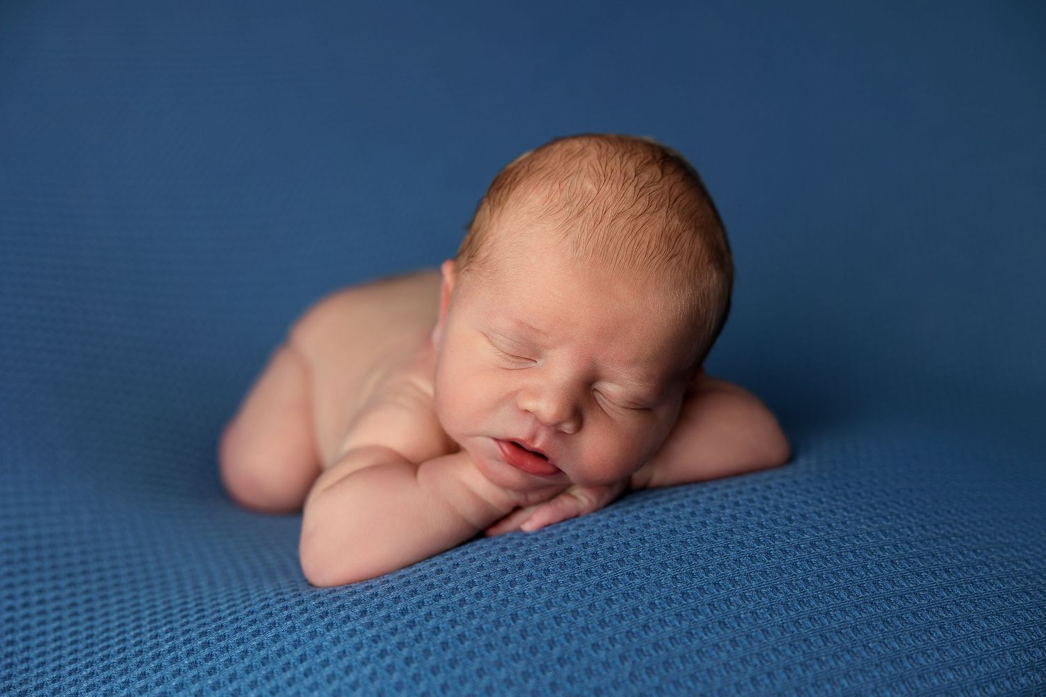 Sleeping newborn baby rests peacefully on textured blue blanket during professional photo session.