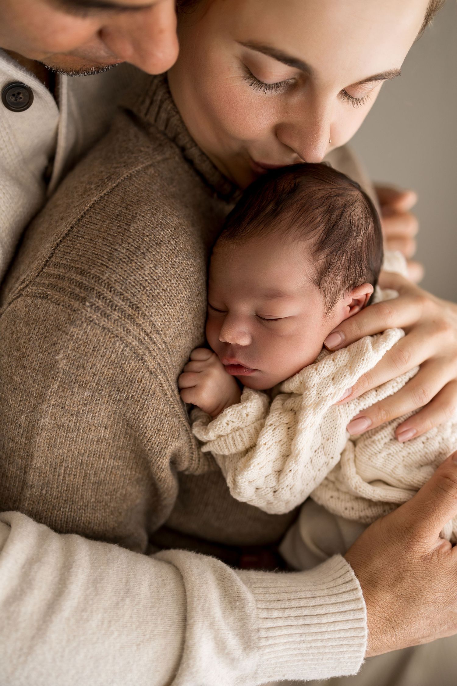 Parents tenderly holding and kissing their sleeping newborn baby wrapped in a white knit blanket.