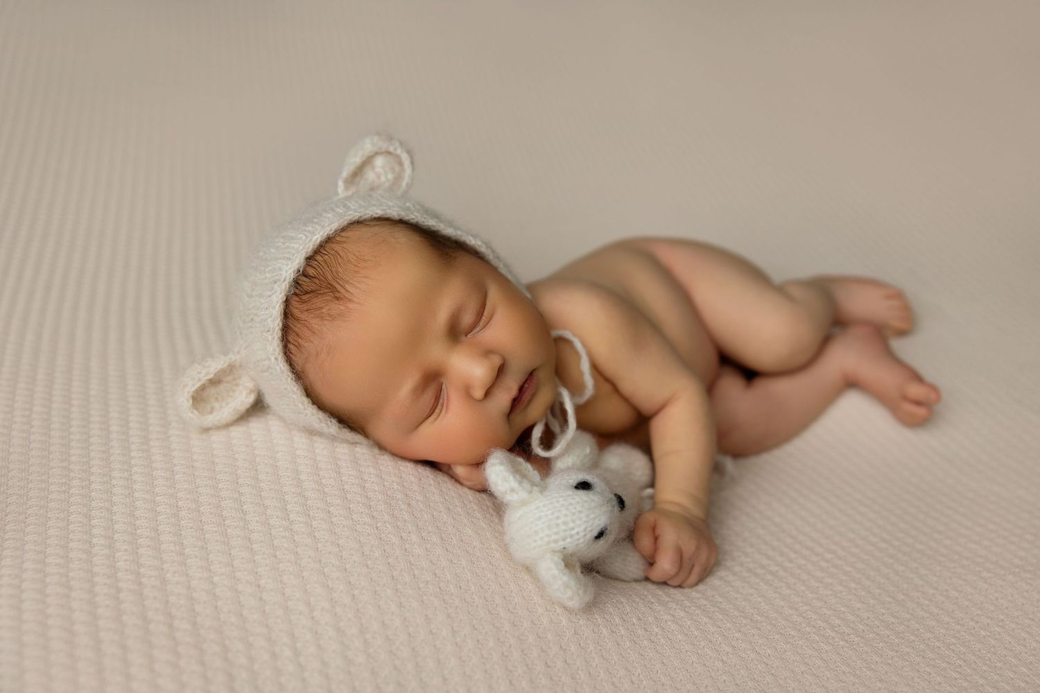 Sleeping newborn with white hat cuddling small toy bunny on neutral backdrop.