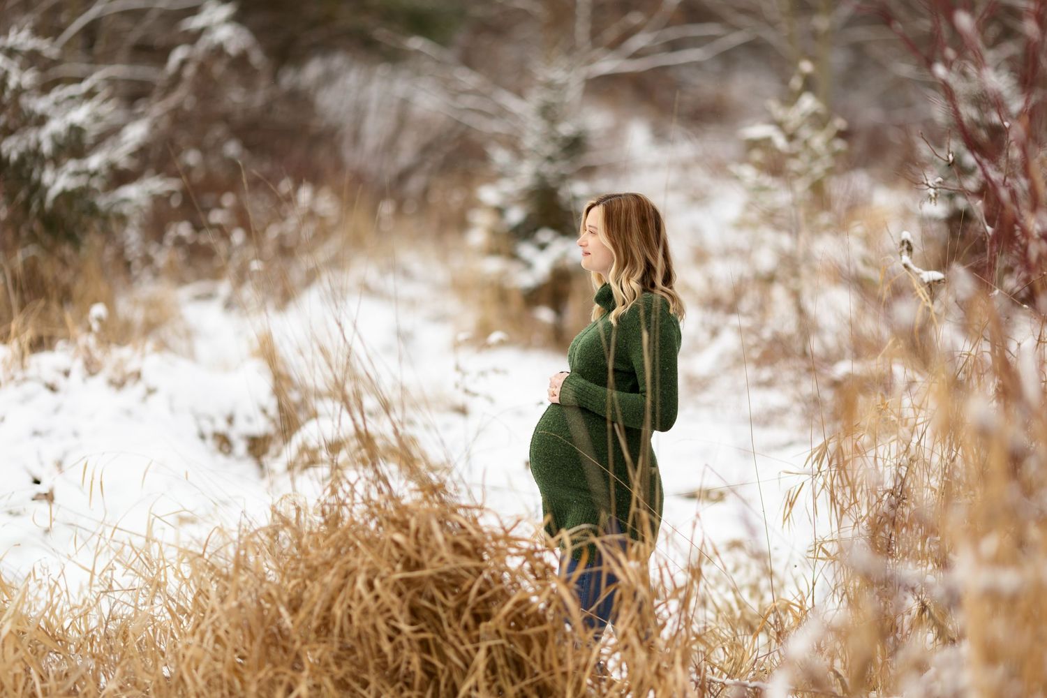 Woman in green sweater poses during winter maternity shoot with snow and dried foliage.