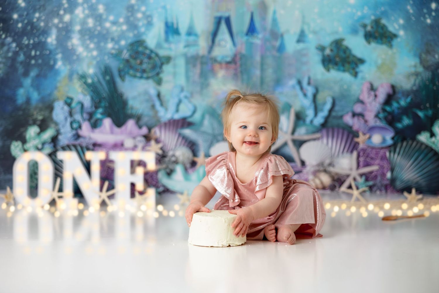 Baby in pink dress sits with birthday cake against magical underwater castle backdrop.