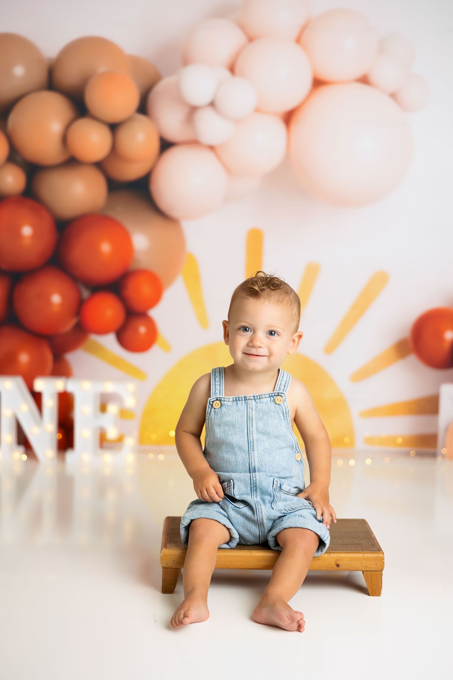 First birthday portrait on wooden stool with balloon and sun-themed backdrop.
