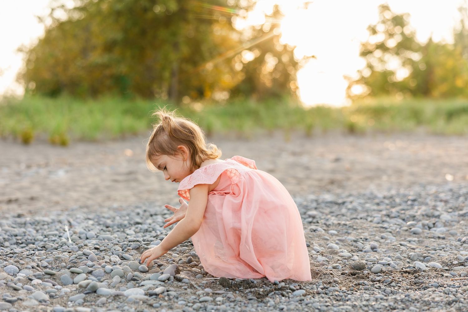 A young child in a pink dress bends down to explore the pebbly ground during golden hour sunset.