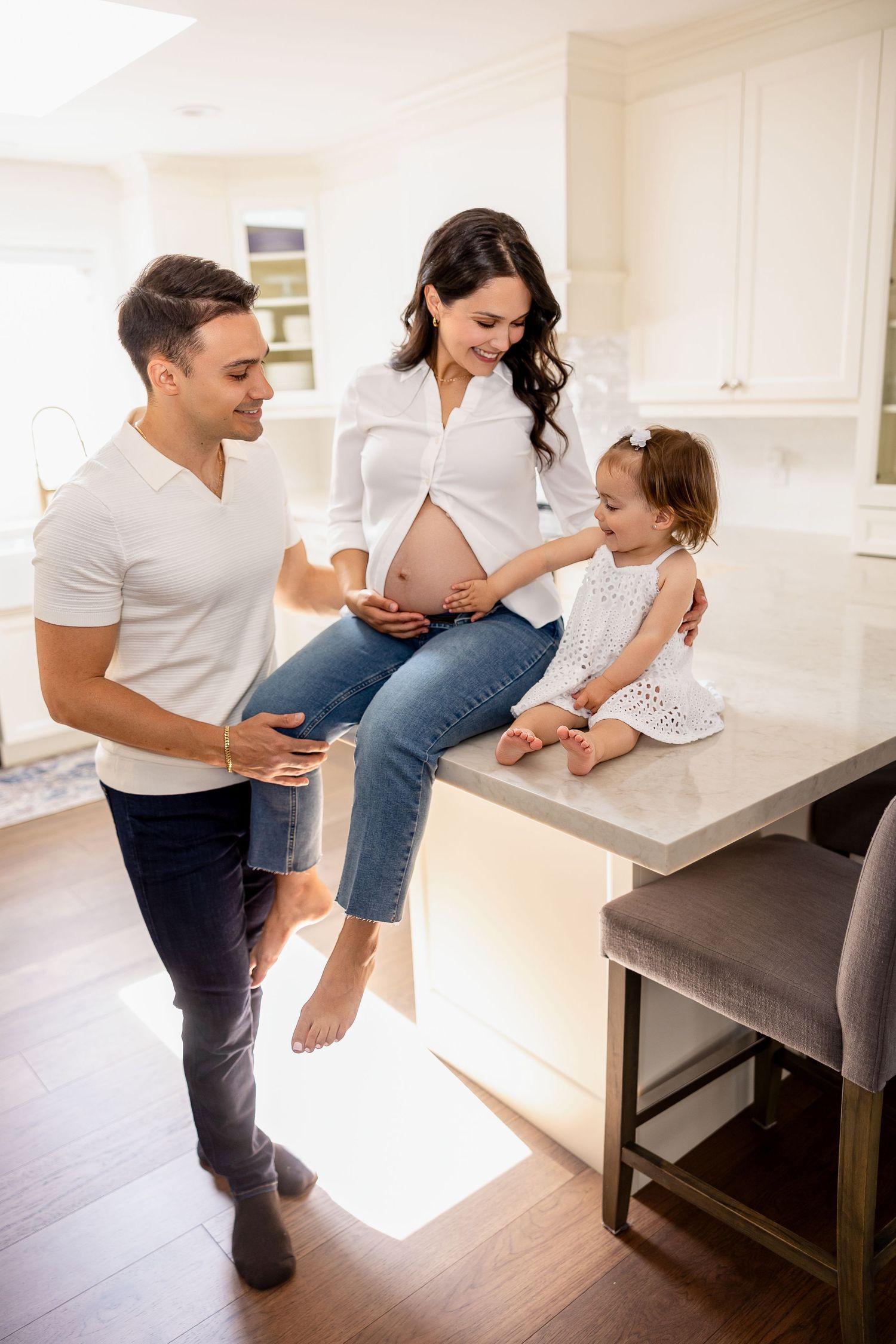 Happy family spending time together in bright modern kitchen as pregnant mother sits on countertop with young daughter.