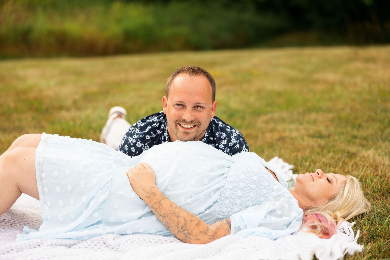 Smiling couple lying together on a white blanket in a grassy outdoor setting during a photo session.