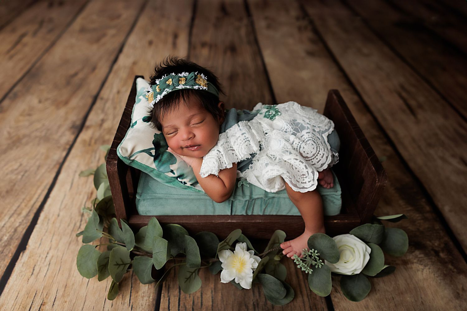 Sleeping newborn posed with greenery and white flowers on wooden floor.