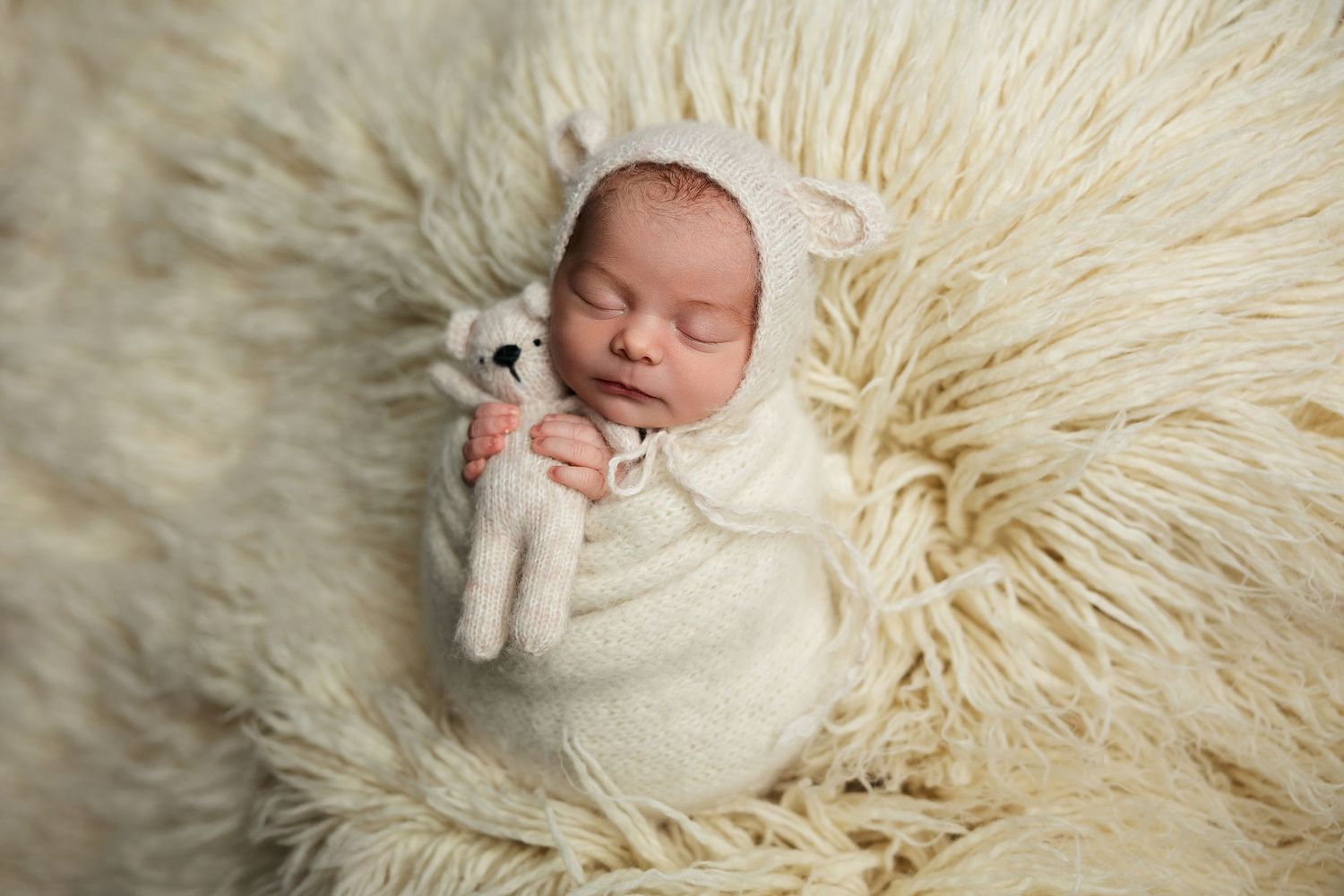Sleeping newborn wrapped in white holding small stuffed toy on cream fluffy rug.