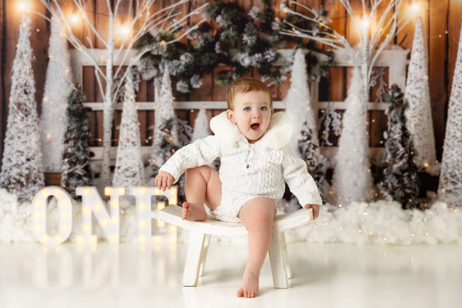 Baby in white sweater sits on chair with winter forest backdrop and illuminated ONE sign.