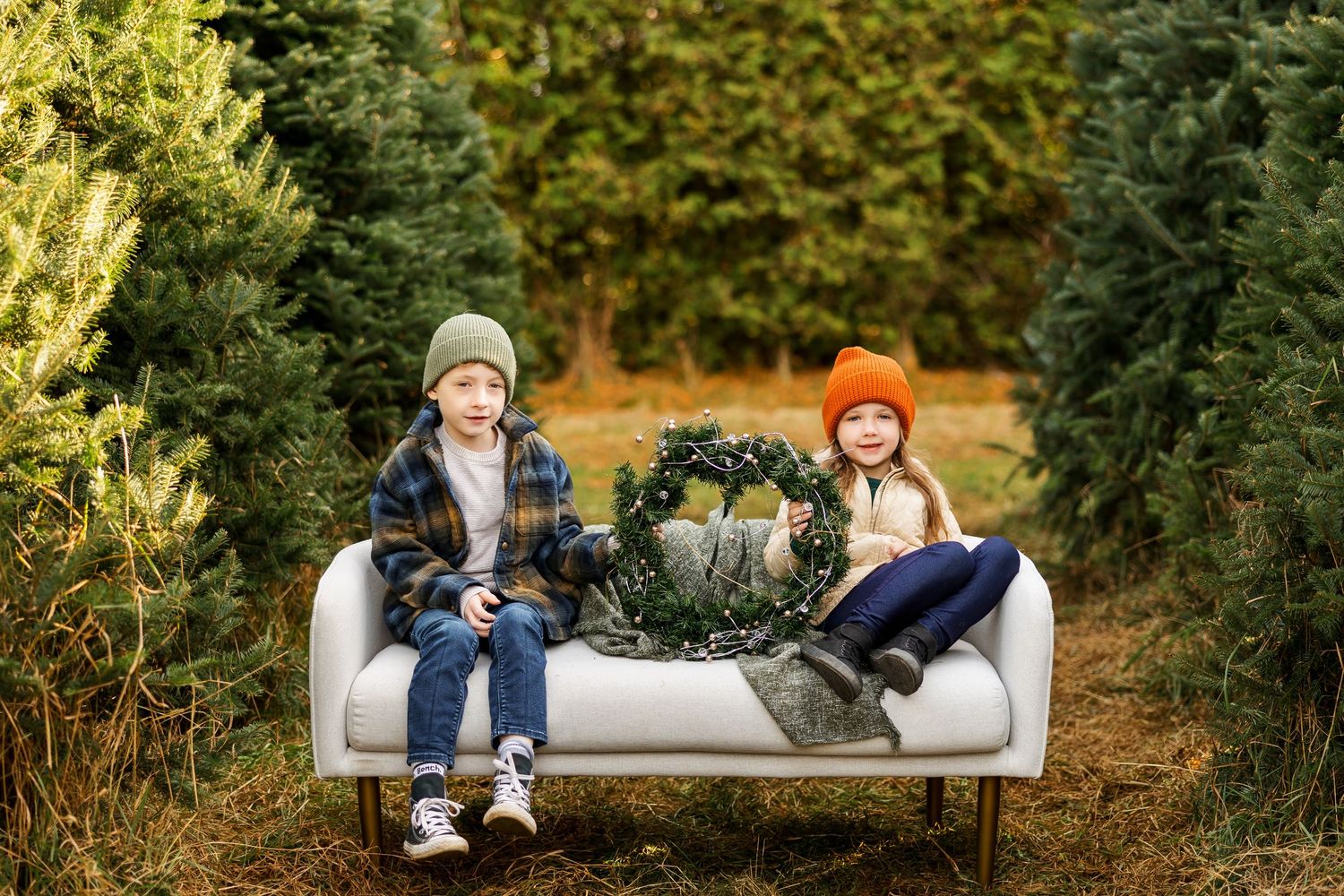 Two kids sitting on a white couch among evergreen Christmas trees with a festive wreath.