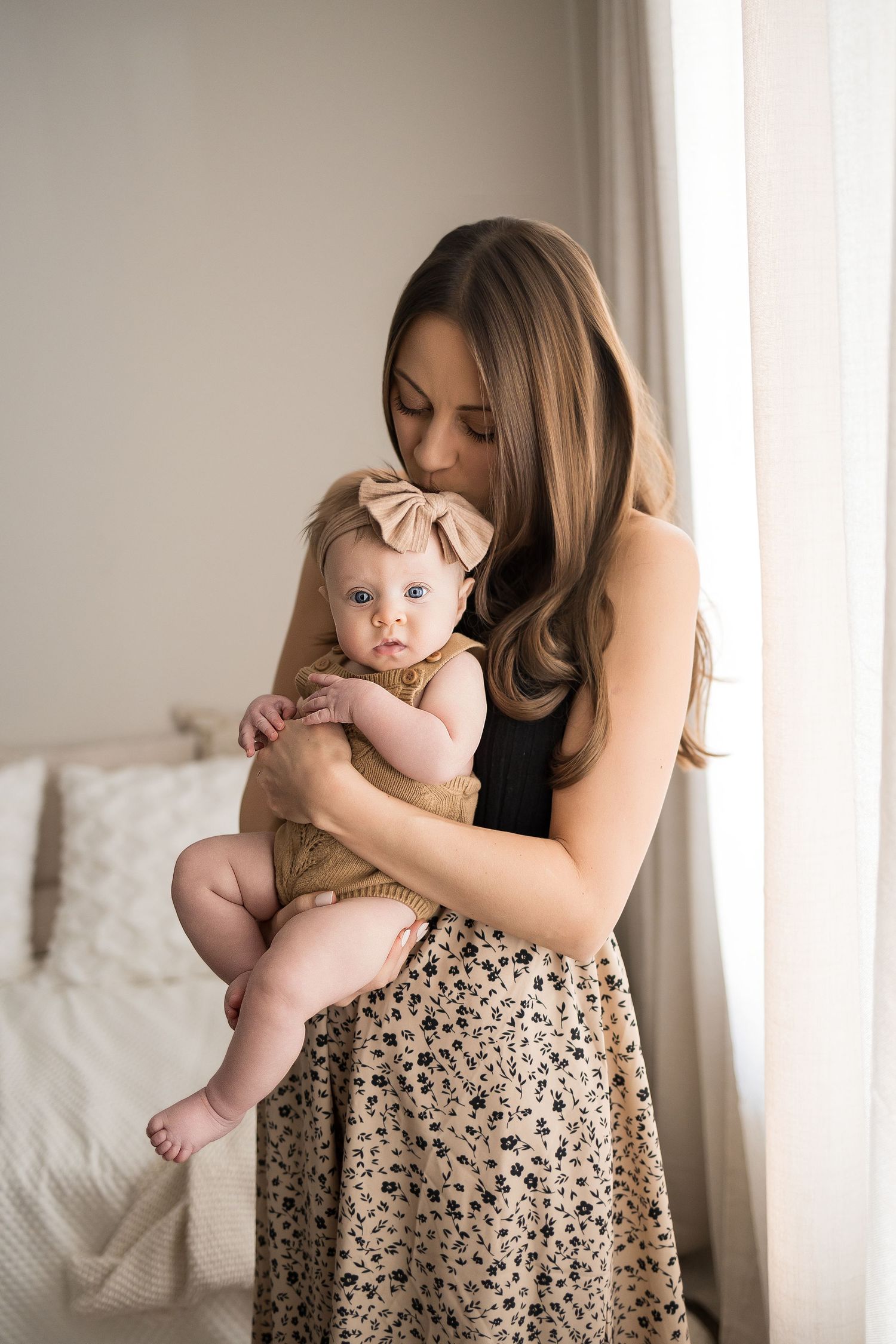 A brunette mother in a floral skirt tenderly kisses her infant baby wearing a tan bow headband.