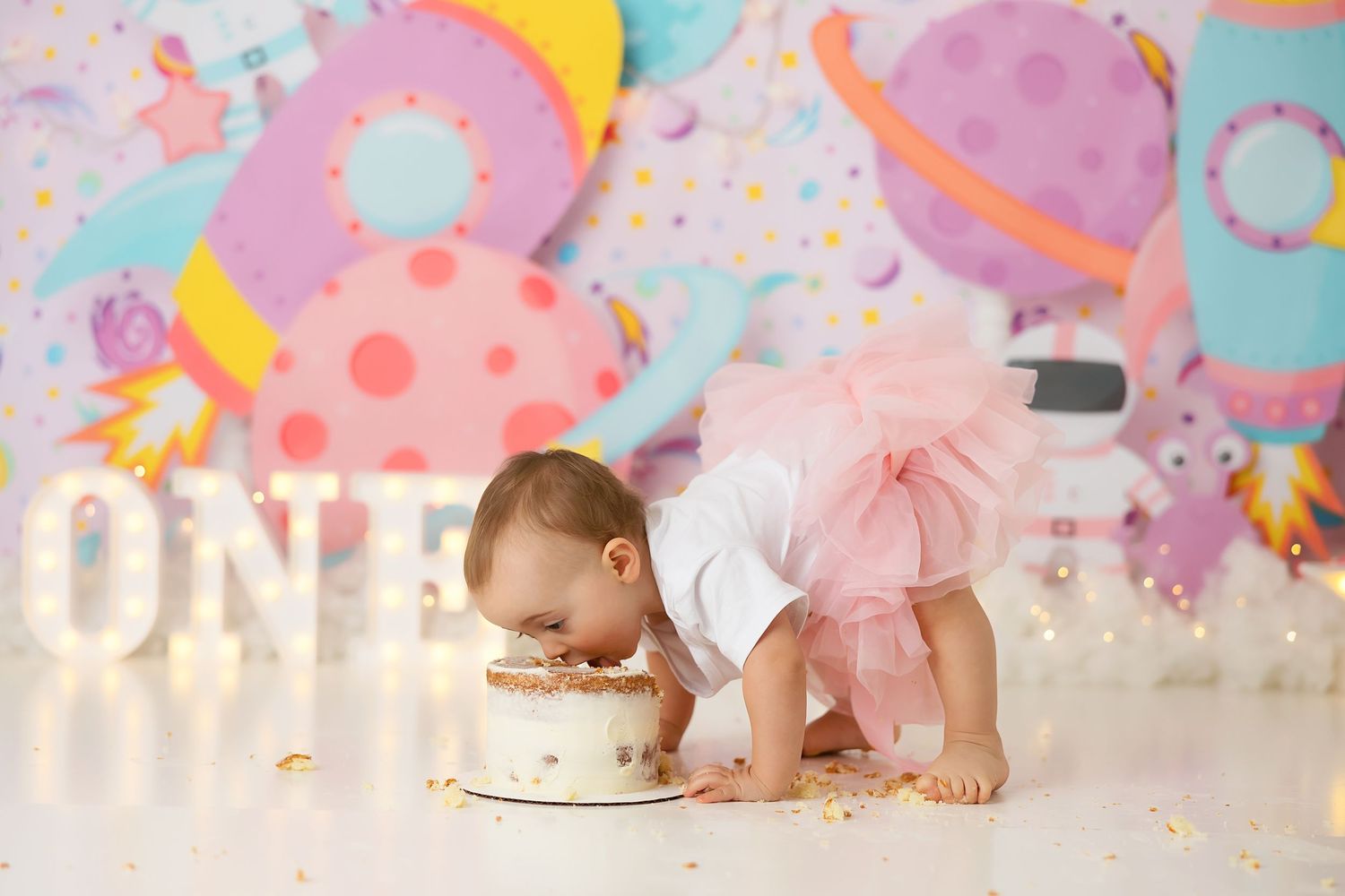 A baby in white clothing leans over to taste a birthday cake against a festive pastel backdrop.