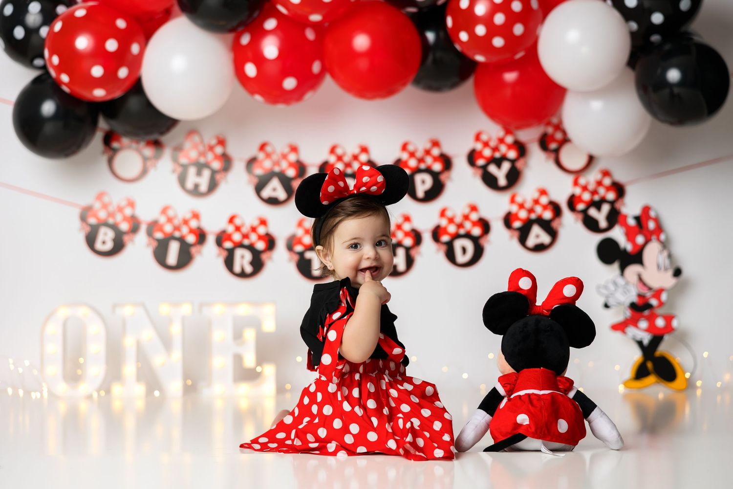 Minnie Mouse themed first birthday setup with red and black balloon arch and illuminated ONE sign.
