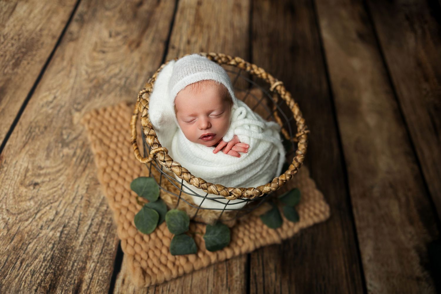 Sleeping newborn wrapped in white swaddle inside wicker basket on wooden surface.