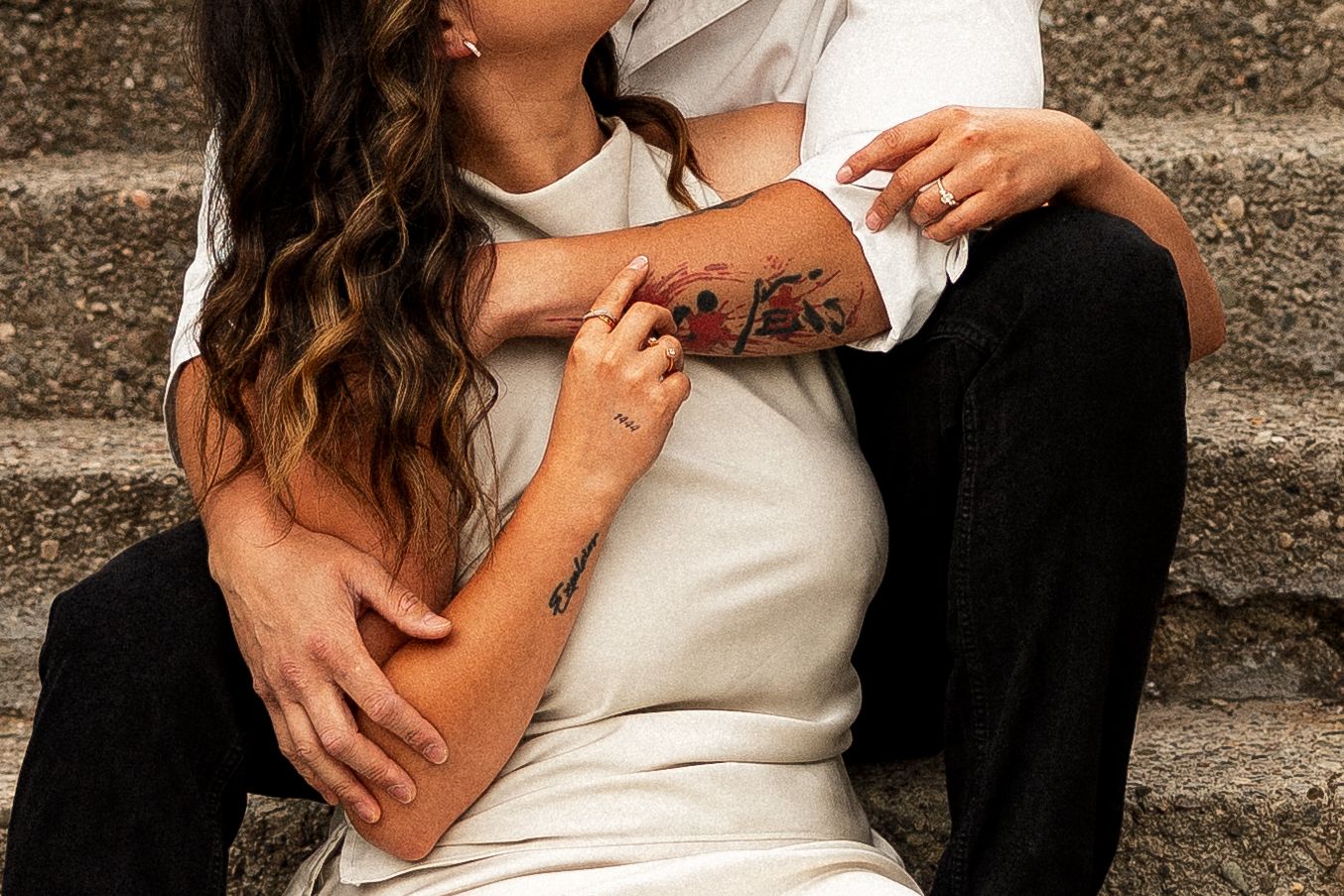 Couple sitting on stone steps embracing, woman with long wavy hair and tattoos, man in white t-shirt and black pants.