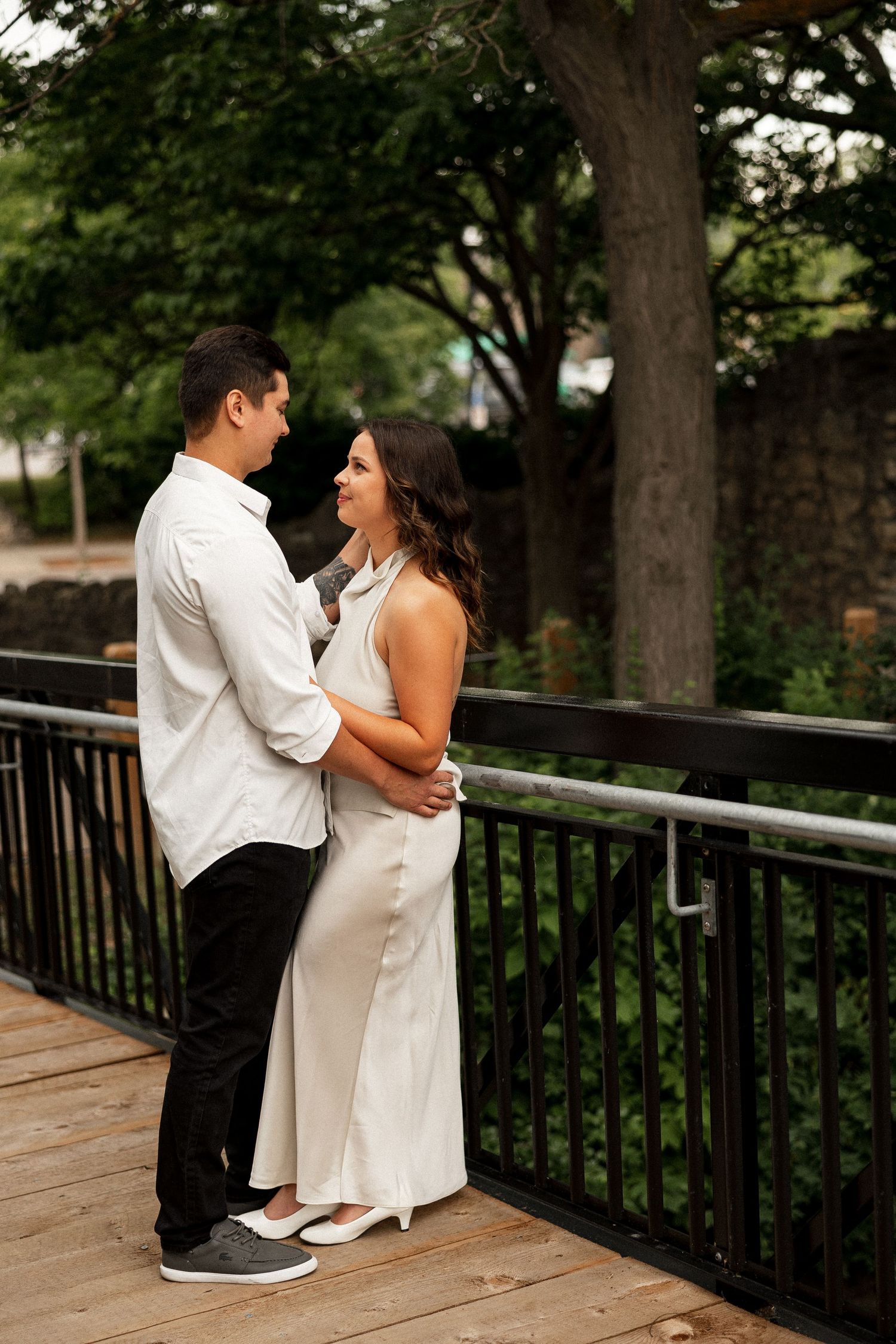 A couple in elegant attire embraces on a wooden bridge surrounded by lush greenery during an engagement session.