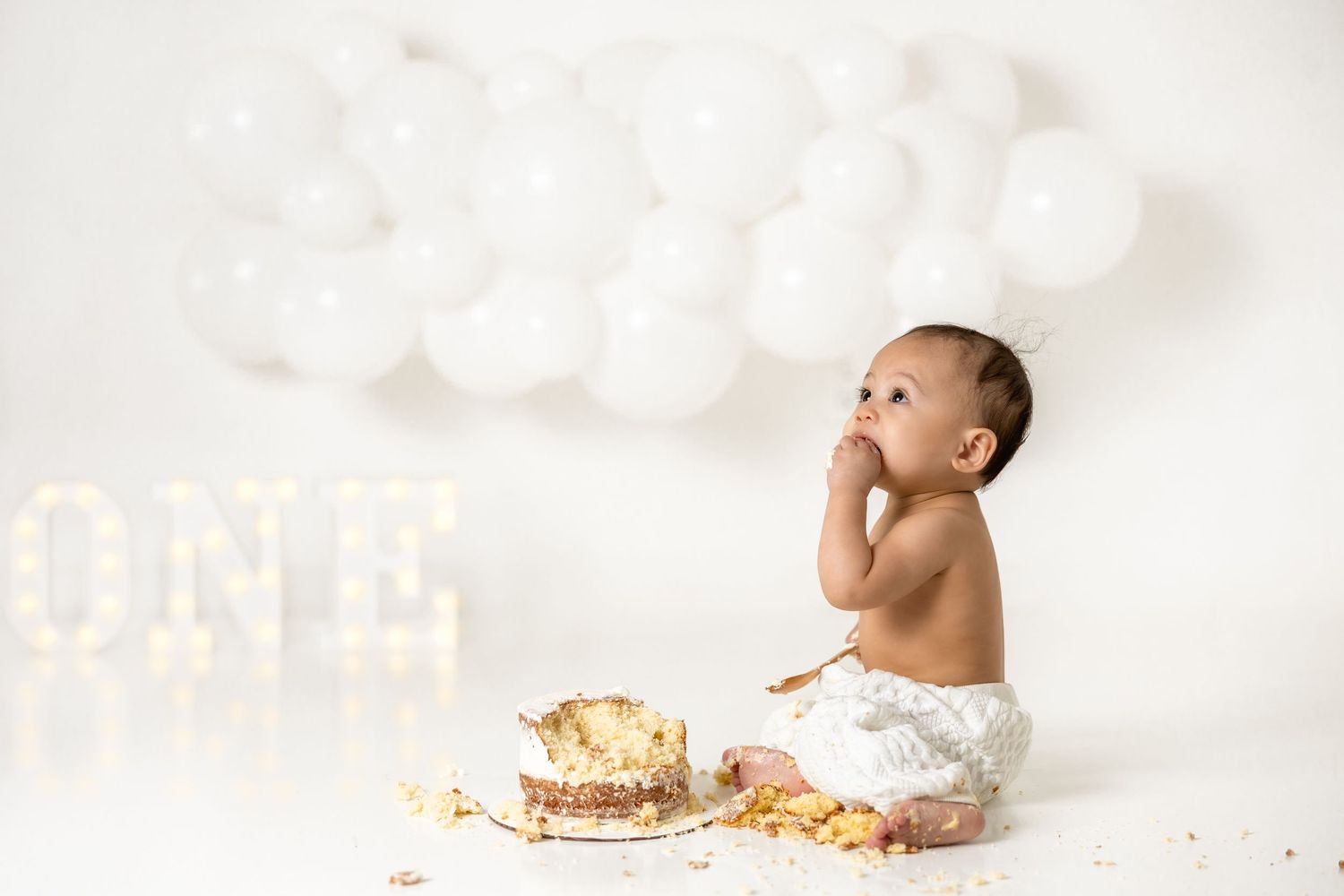Minimalist white cake smash setup with cloud balloons and illuminated ONE sign.