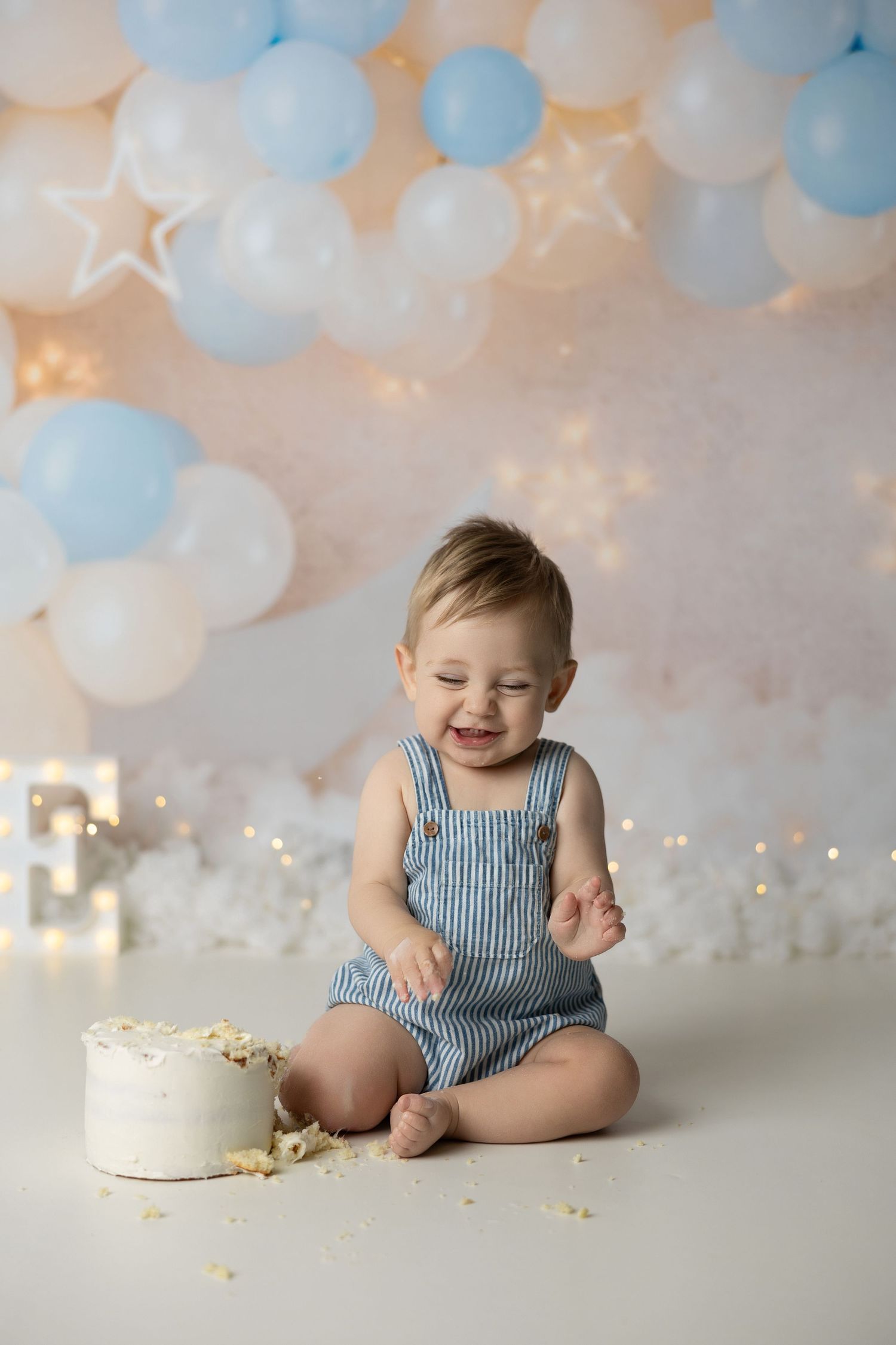 Festive first birthday setup with blue balloons, twinkling lights, and a white smash cake celebration.