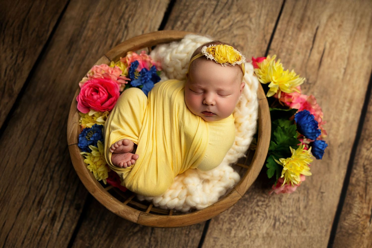 Sleeping newborn wrapped in yellow fabric with colorful flowers in wooden bowl.