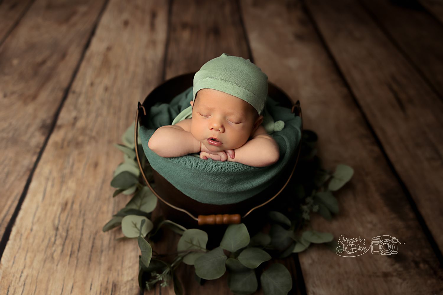 Sleeping newborn in green cap nestled in dark bowl with foliage on wooden surface.