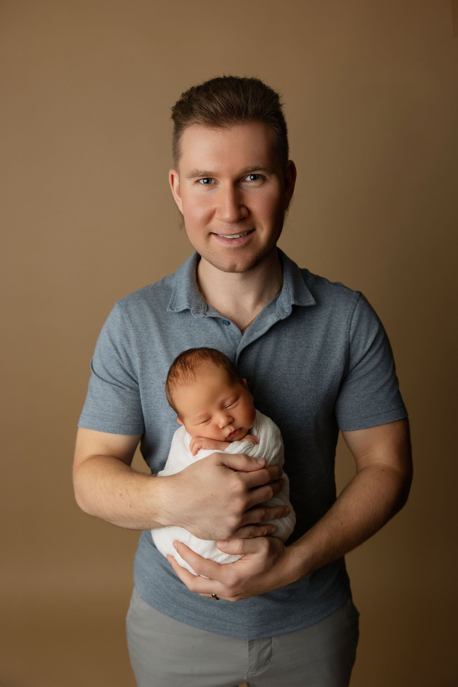 Studio portrait of new parents with sleeping infant against brown backdrop.