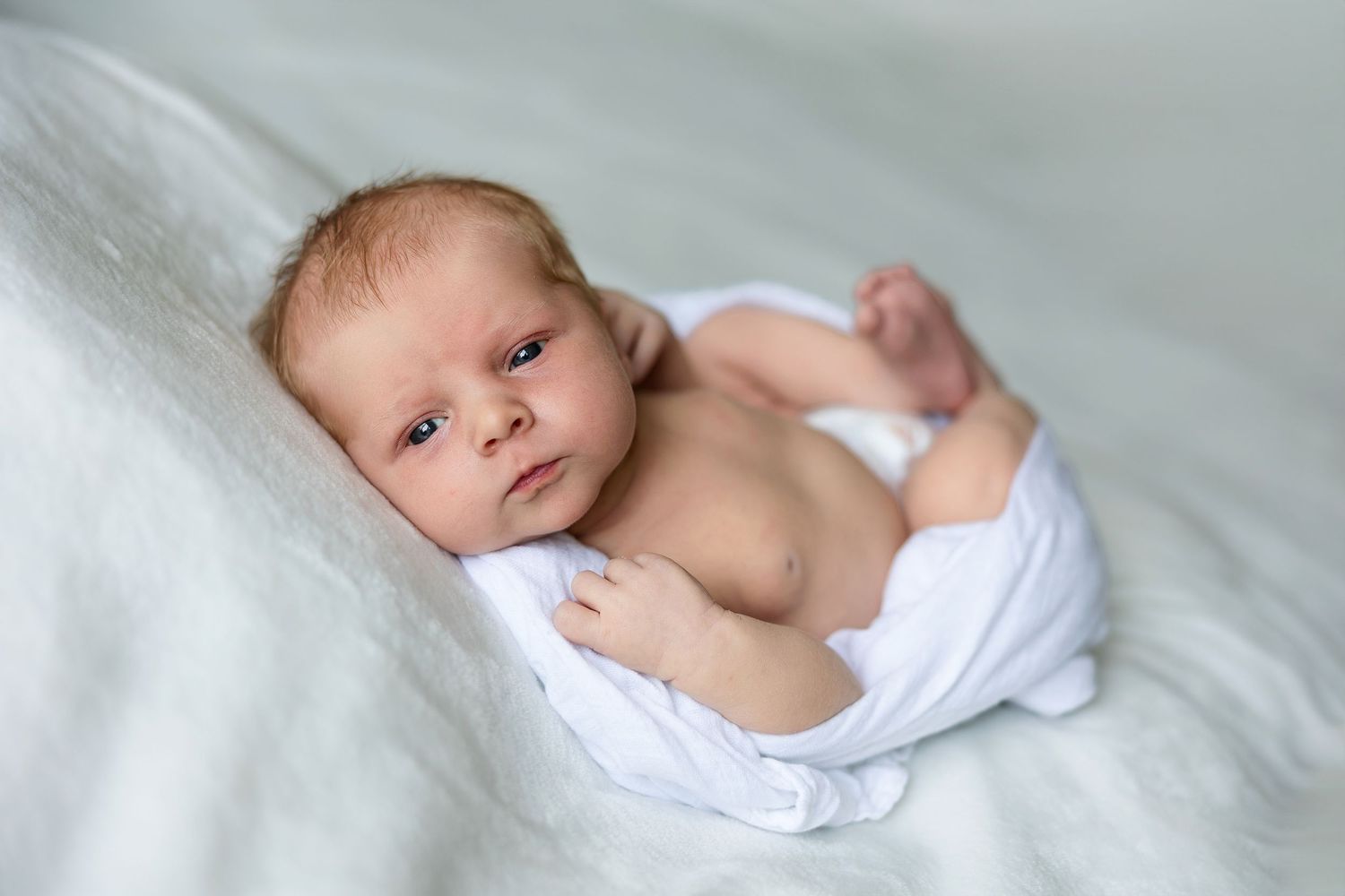 Newborn baby wrapped in soft white blanket laying on light fabric background.