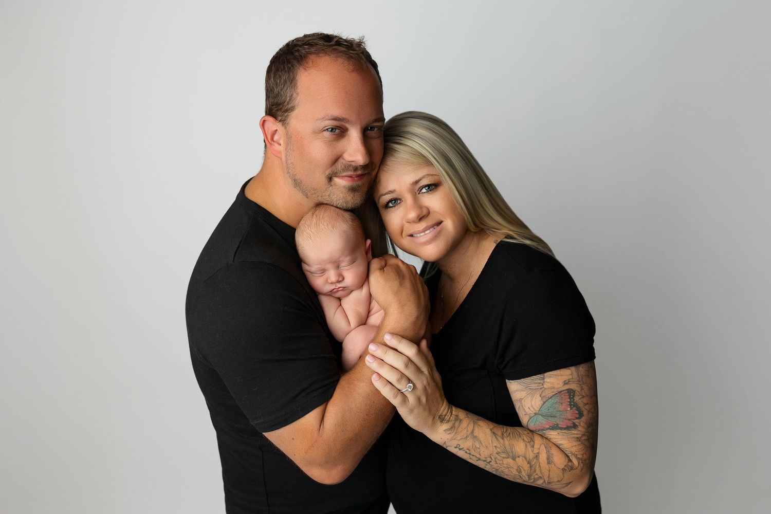 A family portrait against a gray background shows parents in black clothing holding their newborn baby.