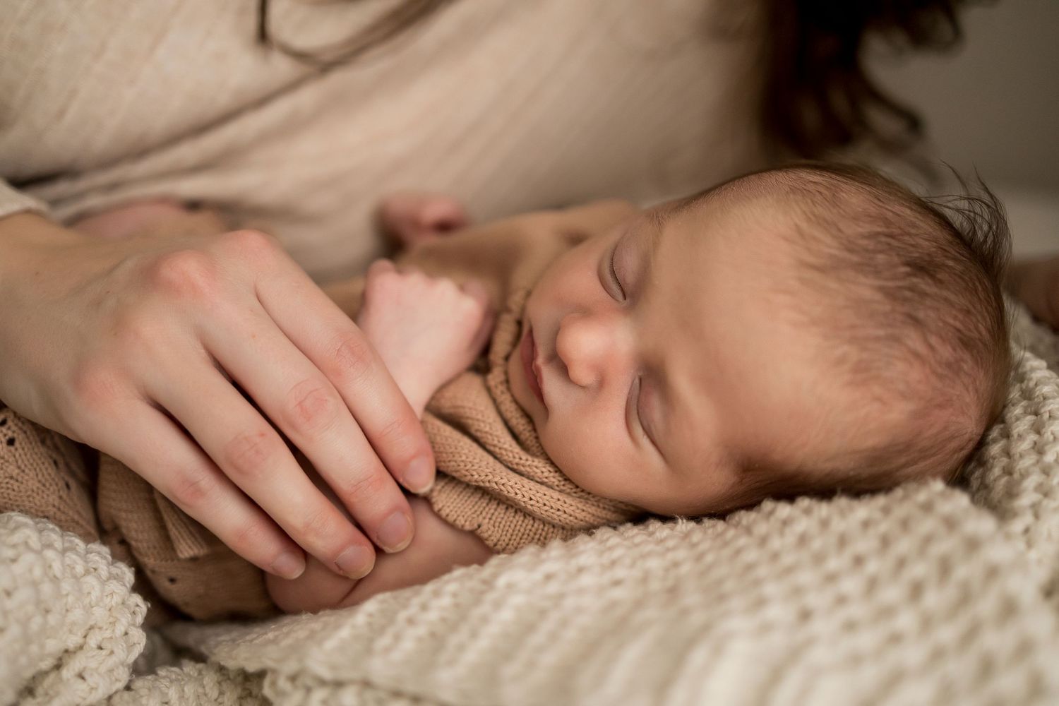 Sleeping newborn baby wrapped in beige knit blanket rests peacefully on textured cream bedding.
