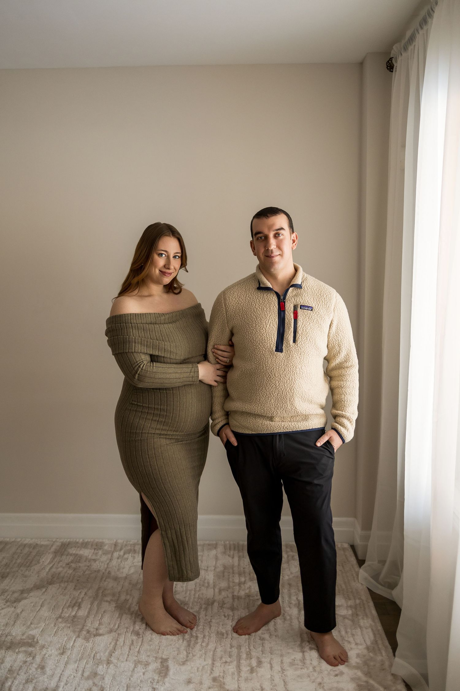 A couple in coordinating neutral sweaters poses together against a white wall in a minimalist setting.