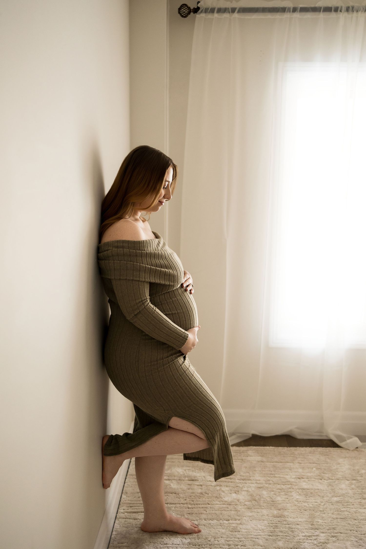 Silhouette of an expectant mother in a cozy knit dress standing by a sunlit window.