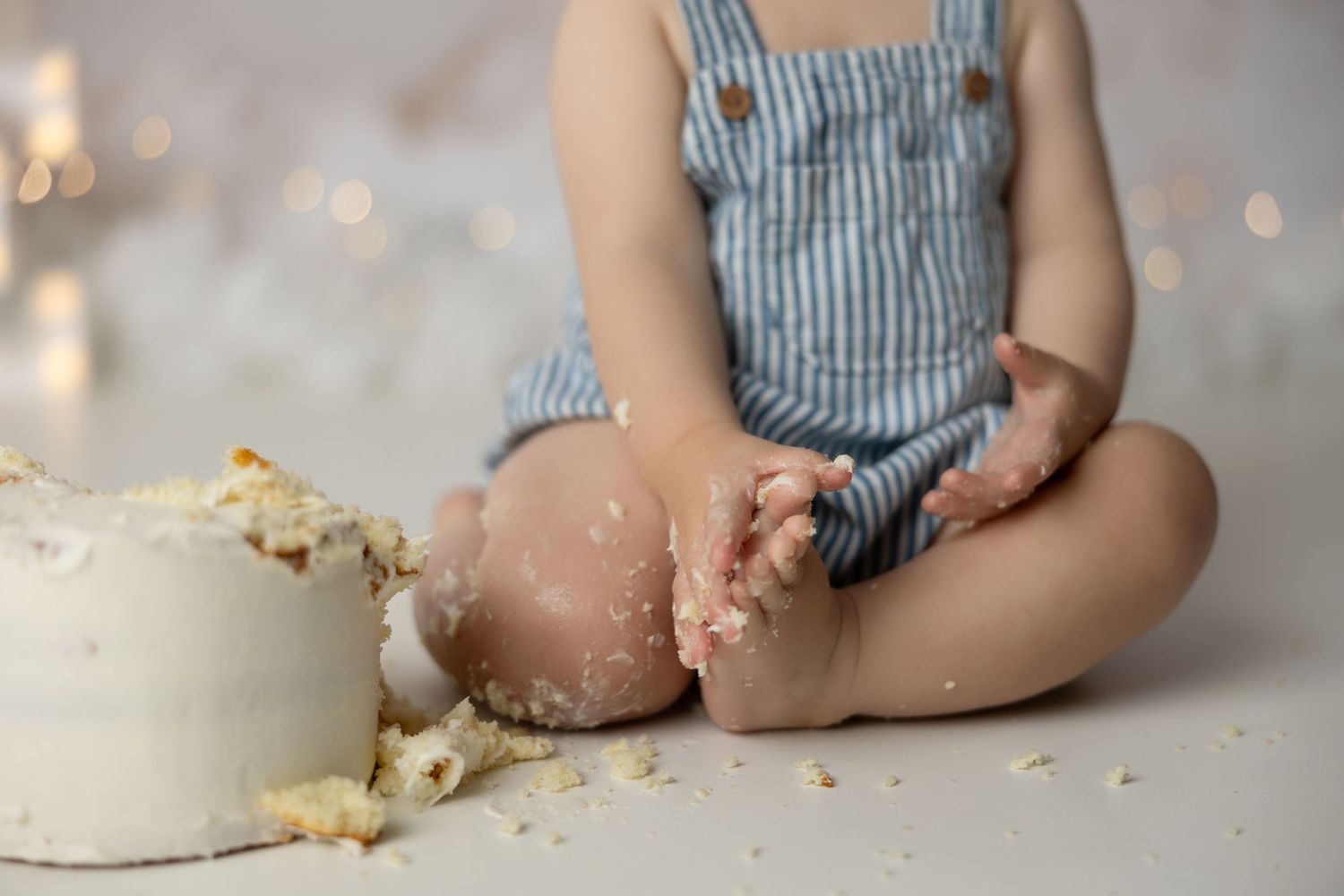 Close up of small child in striped outfit sitting with smashed cake and crumbs on white floor.