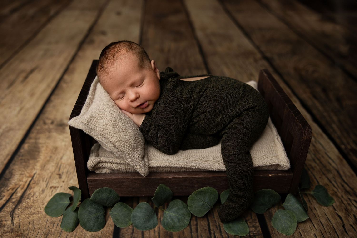Newborn sleeping peacefully in wooden box with eucalyptus leaves on rustic wooden floor.