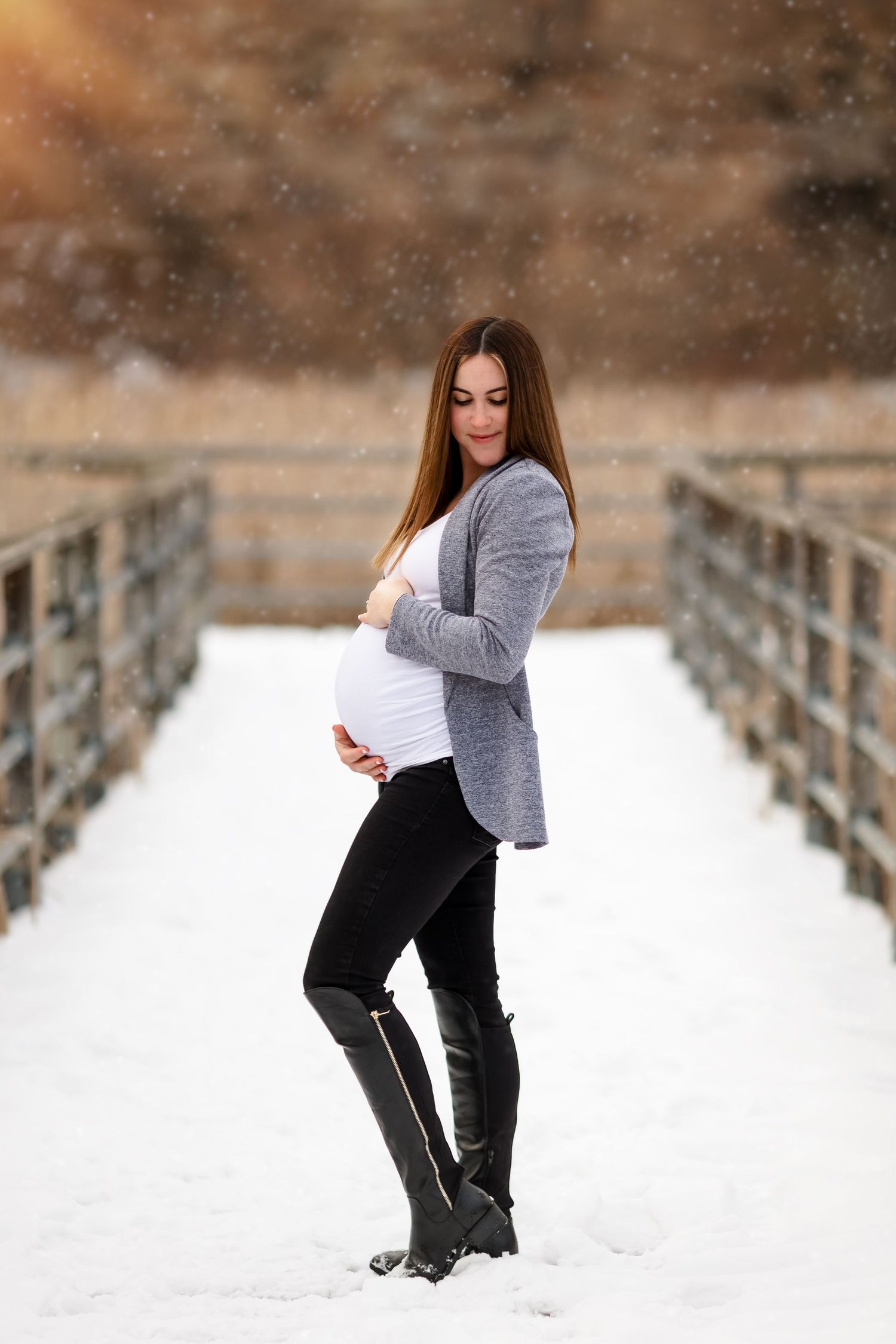 A maternity photo of an expectant parent in a gray sweater and black boots standing on a snowy bridge.