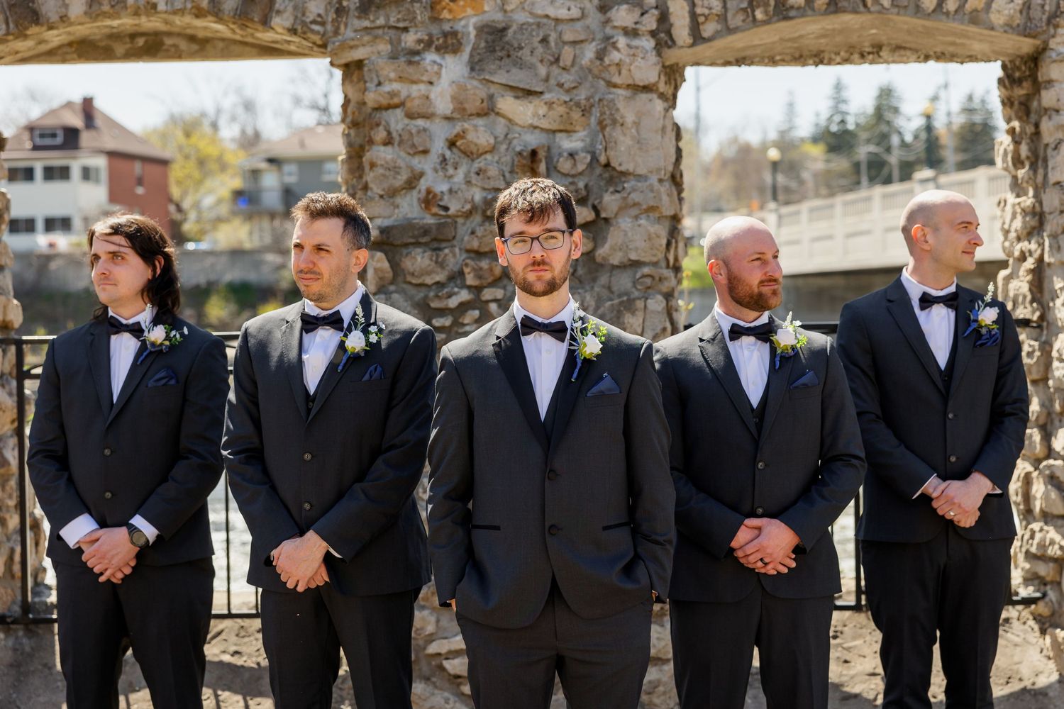 Groomsmen in black tuxedos and bow ties lined up against stone wall backdrop.