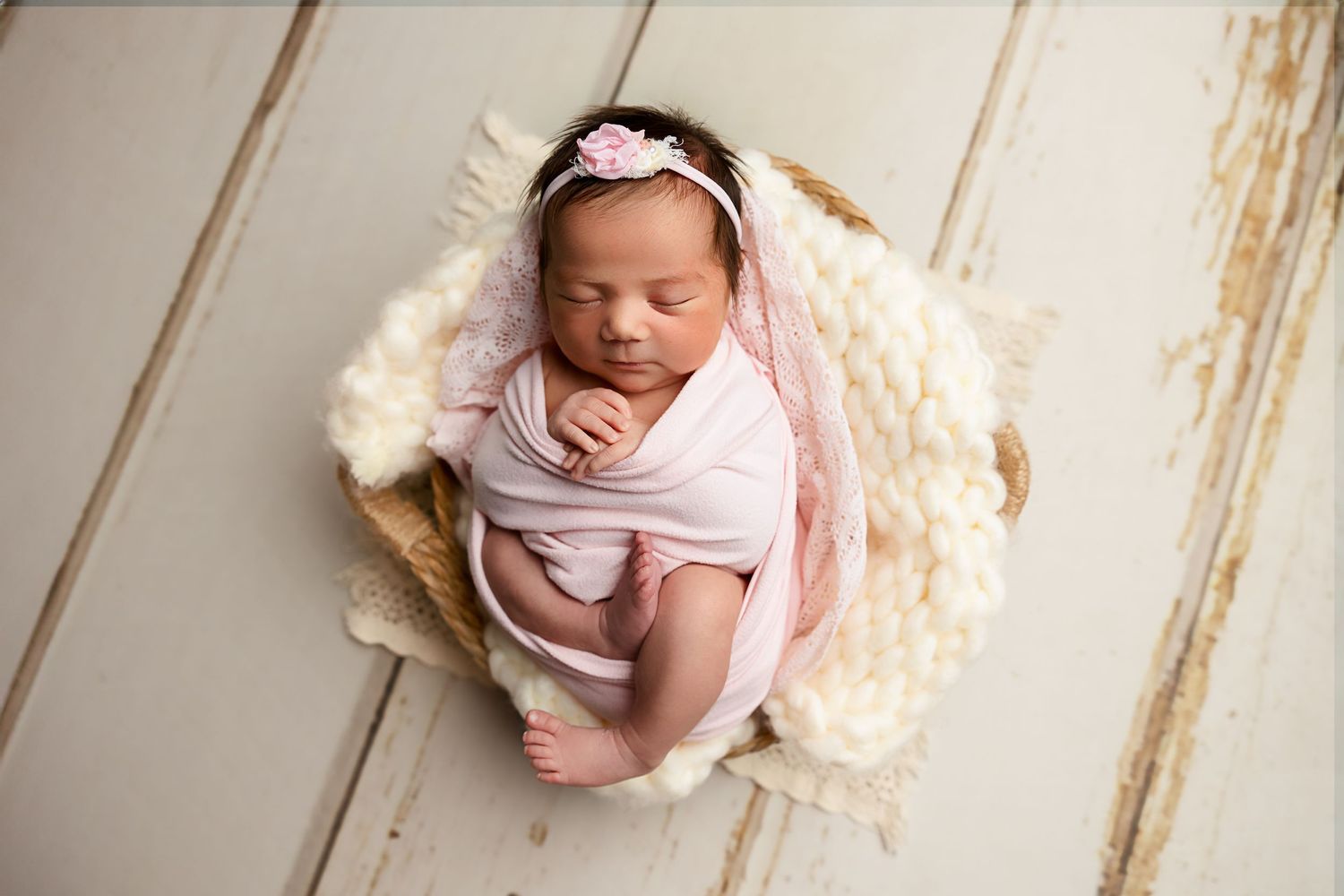 Peaceful newborn swaddled in soft pink fabric sleeping in round basket with fluffy cream blanket.