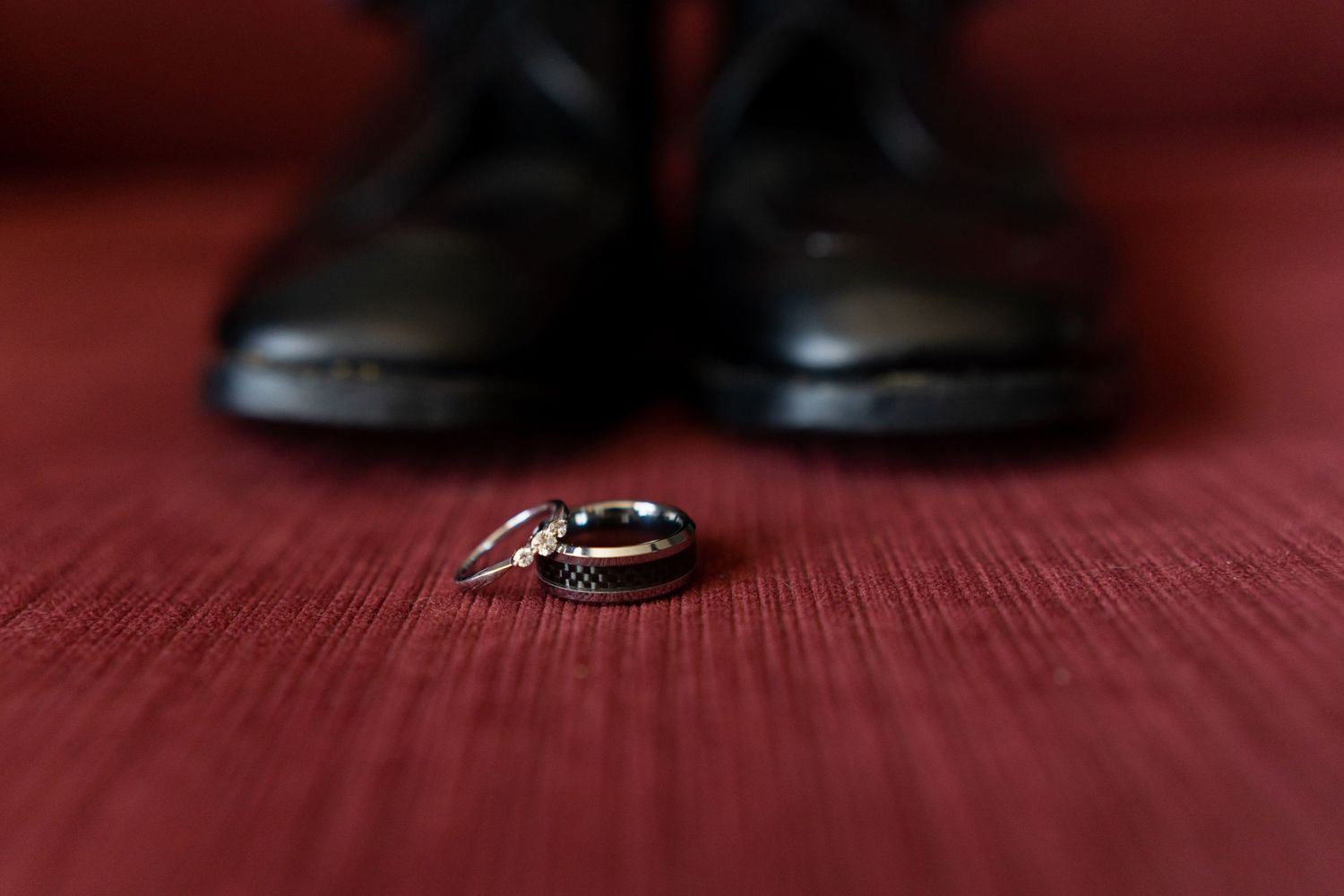 Wedding rings and dress shoes arranged on a red carpet surface with dramatic lighting and shallow depth of field.