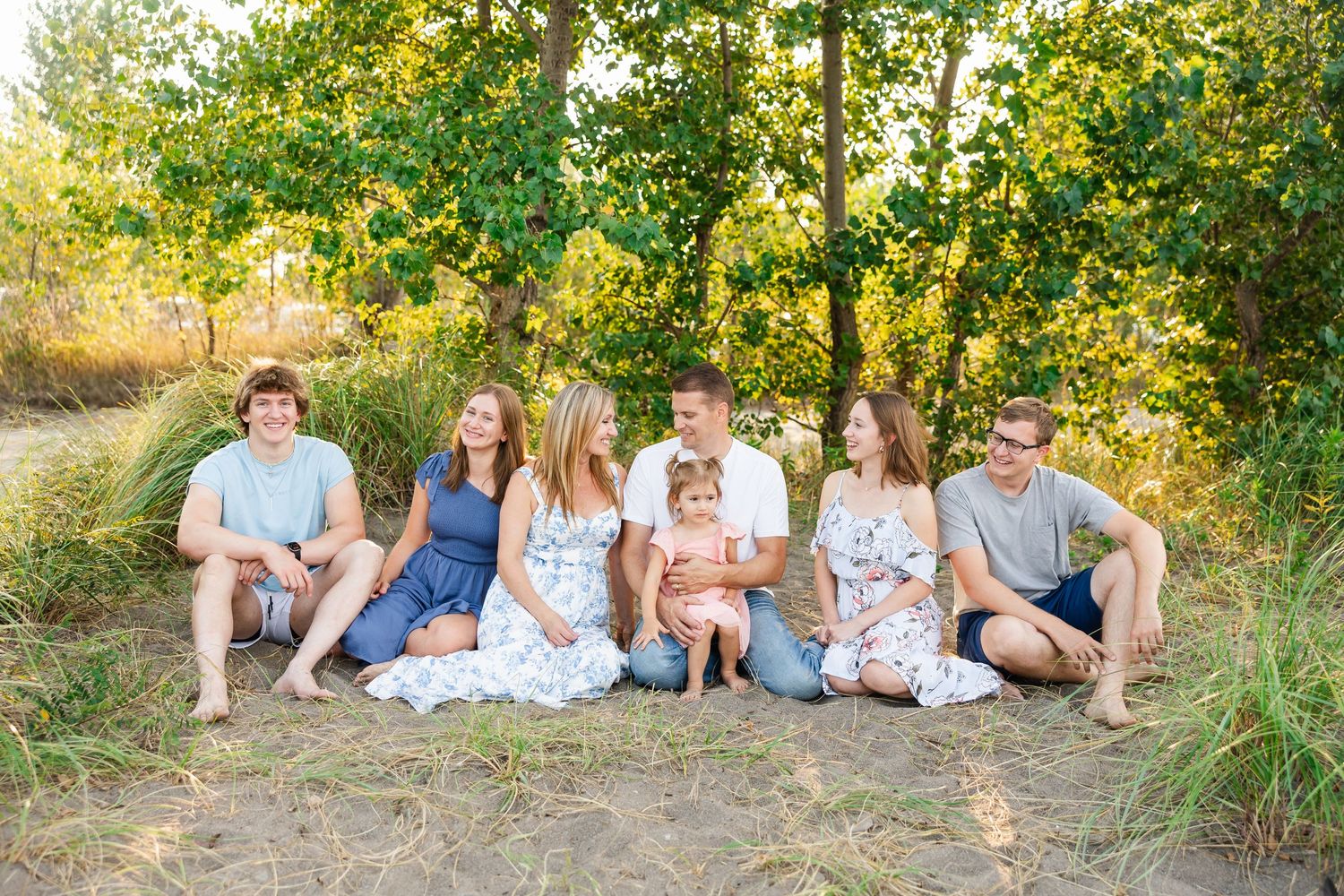 A group sits together outdoors on a grassy hillside with trees and golden sunlight in the background.