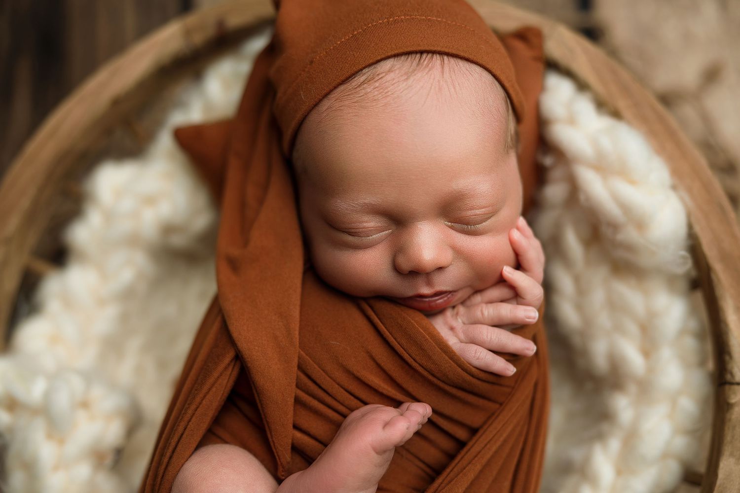 Sleeping newborn baby wrapped in a rust-orange swaddle with matching bear hat, nestled in a wooden bowl.