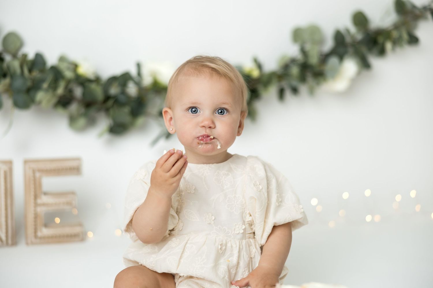 Baby girl in cream romper eating cake smash treat, sitting against white backdrop with greenery garland and fairy lights.