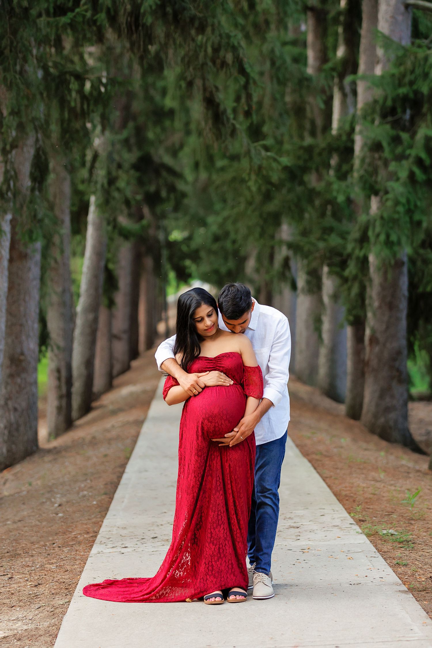 Maternity photoshoot of couple on tree-lined path with woman in flowing red dress.