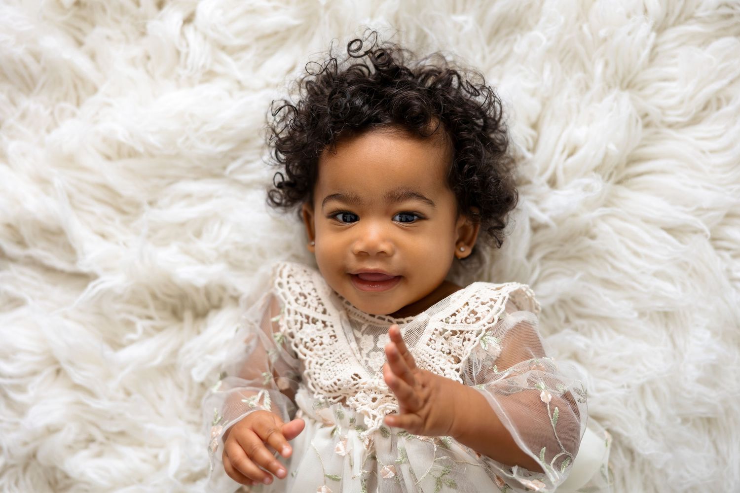 Baby in white lace dress poses on fluffy white rug.