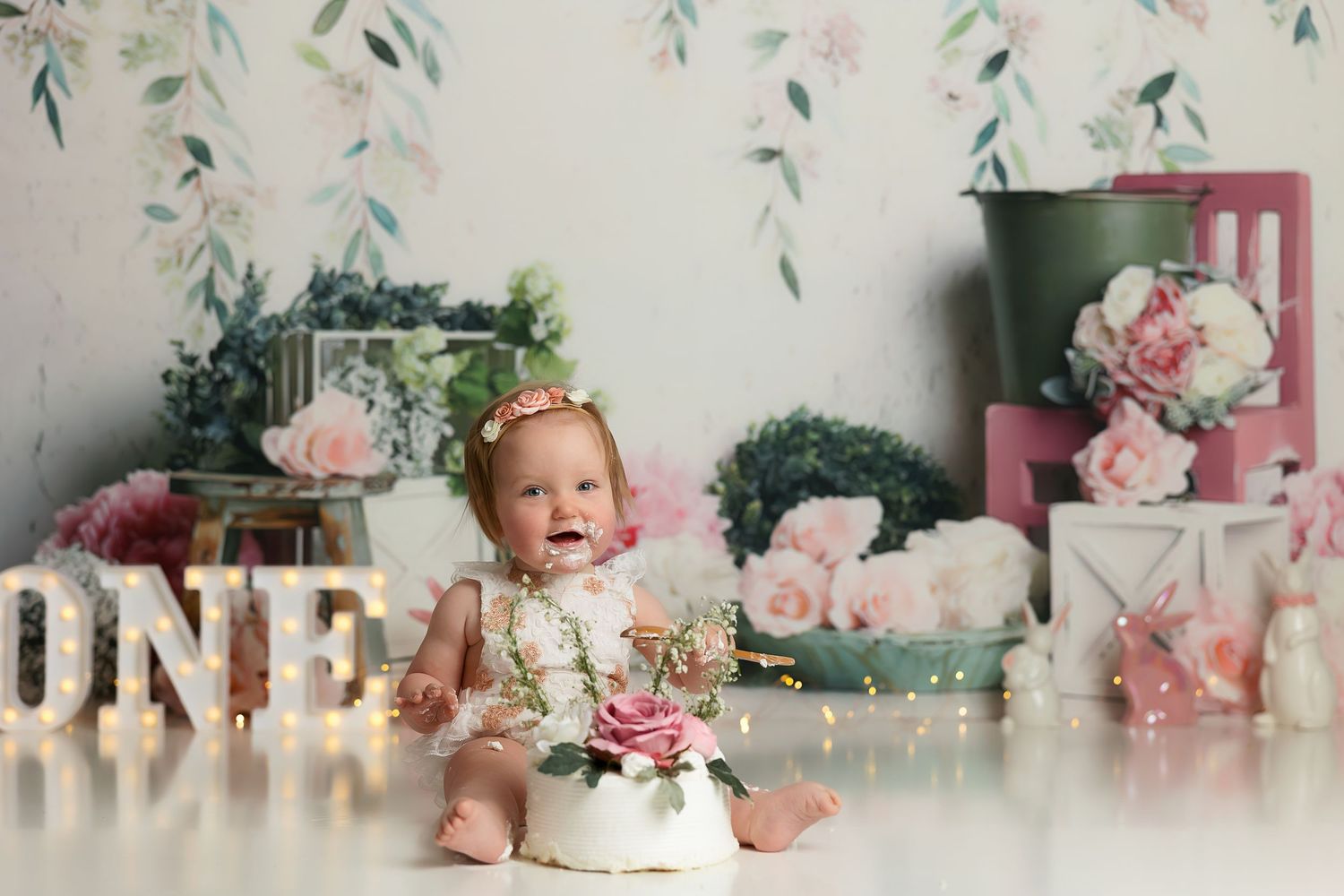 Floral themed first birthday setup with greenery, illuminated ONE sign and vintage decor elements.