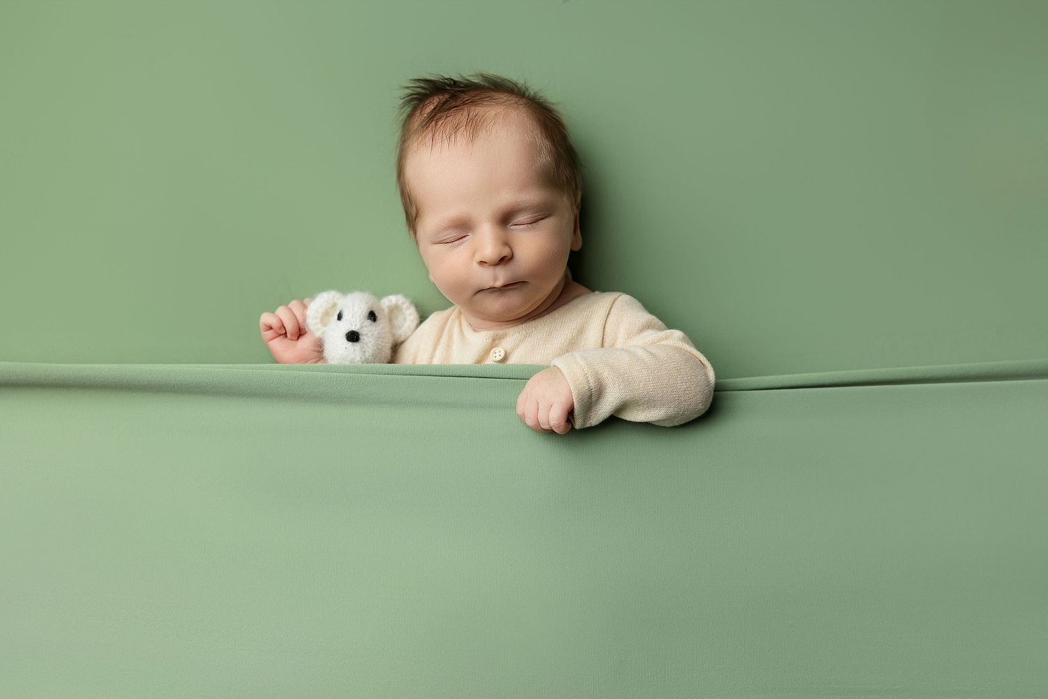 Baby in cream outfit resting on mint green backdrop with small toy.