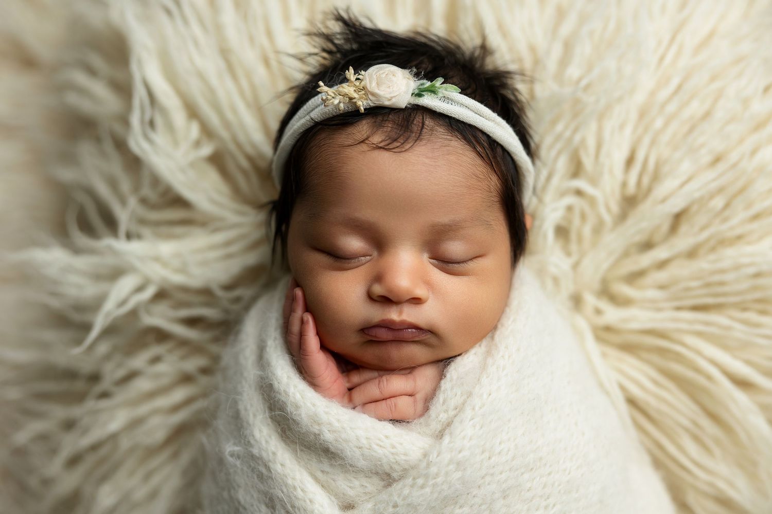 Peaceful newborn wrapped in white swaddle sleeping on fluffy cream backdrop.