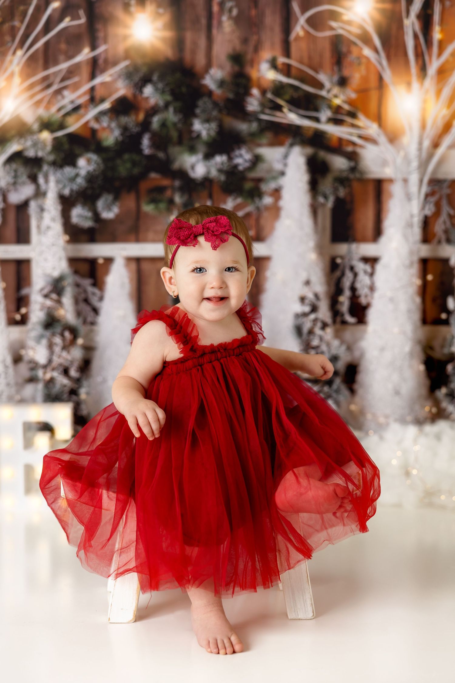 Baby in red tulle dress sits on white chair against snowy winter wonderland backdrop.