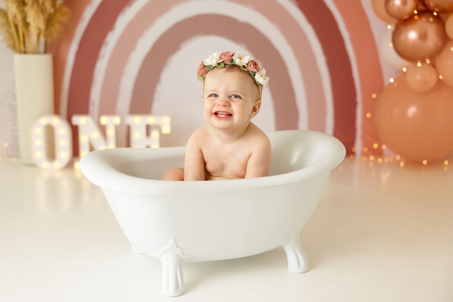 Classic first birthday bath tub photo with peach balloon arch and rainbow backdrop.