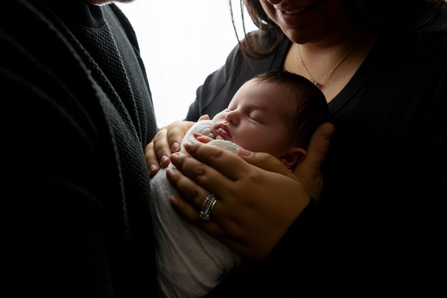 Intimate family portrait of parents cradling newborn in dramatic lighting against white background.
